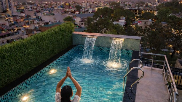 Una persona en una piscina con vistas al atardecer, una cascada y un vaso de zumo de naranja al borde.