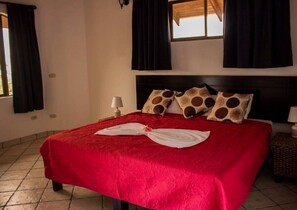 A hotel room with a bed, a window with curtains, a bedside lamp, and a small table with a lamp. - Jaco Castle by Rey (Jacó)