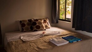 Ein Bett mit einer beige farbenen Bettdecke, zwei Kissen mit einem Rundmuster, ein weißes Handtuch und eine blaue Tasche auf dem Bett.