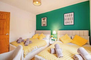 Une chambre d’hôtel avec deux lits, chacun agrémenté de draps à motifs jaunes et blancs, un mur d’accent vert, une pancarte indiquant « Seules les bonnes ondes sont les bienvenues » et une porte en bois.