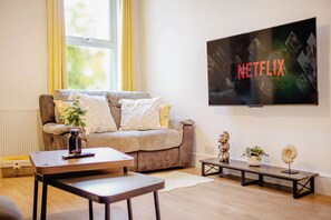 Una sala de estar con un sofá, una mesa de centro, un televisor montado en la pared y una mesita con objetos decorativos.