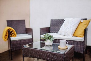Un canapé en osier avec des coussins, une table basse en verre ornée d’une plante et d’une tasse, et un plaid jaune jeté sur une chaise.