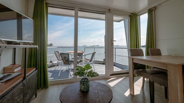 Una habitación con un amplio ventanal corredera que da a un balcón con vistas al agua. Hay una mesa redonda con una planta en el centro de la habitación.