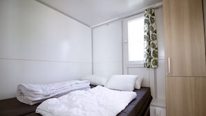 Una cama individual con ropa de cama blanca, una ventana con cortinas de estampado de hojas y un armario de madera.