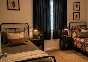 Ein Schlafzimmer mit zwei Betten, einem Fenster mit Vorhängen, Nachttischen mit Lampen und gerahmten Bildern an der Wand.