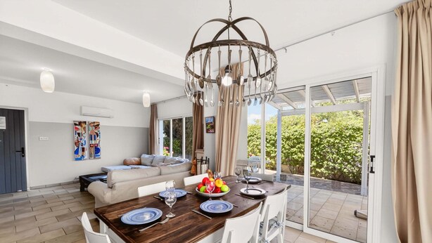 Un comedor con una mesa de madera dispuesta para cuatro, sillas blancas, una araña y vistas a zonas verdes al exterior.