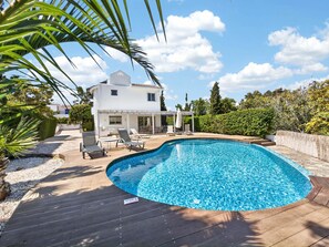 A swimming pool with a wooden deck, a white house with a covered patio, and surrounding greenery. - Villa Marilena: Spacious Protaras Villa with Pool (Protaras)
