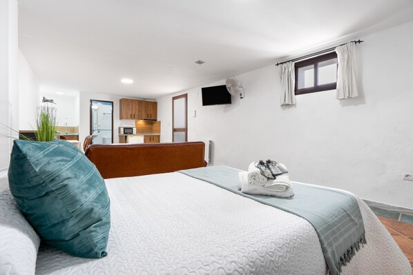 Une chambre à coucher avec un lit, un oreiller, une télévision, une fenêtre et, au fond, un coin cuisine.