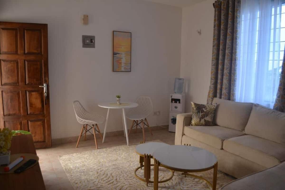 Uma sala de estar com sofá, uma mesa redonda, duas cadeiras, uma porta e um quadro na parede.