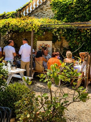 An outdoor gathering with people seated at tables, surrounded by greenery and a stone wall. - Lemon Trees · Courtyard with Flowers and a Pool (Saint-Montan)