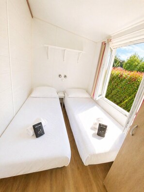 A compact room with two beds, a window with a view of greenery, and a white wall with a shelf. - Cottage Champêtre n°1 à Angliers - 6p (Angliers)