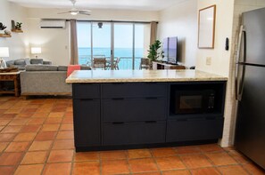 A kitchen with a large island, a built-in oven, and a refrigerator. There is a dining area with a table and chairs, and a view of the ocean through a sliding door. - East End Escape Top-Floor Caribbean Views • Pool • Near Shoy Beach & Buck Island (Christiansted)
