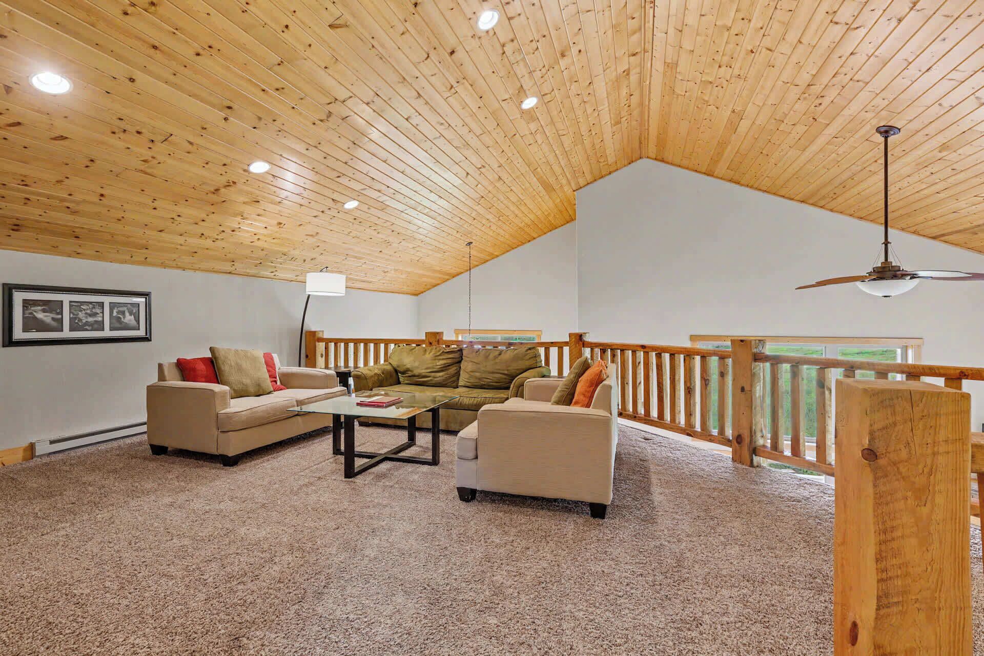 A spacious living room with a high wooden ceiling, a glass coffee table, two sofas, and a wooden railing overlooking a view.