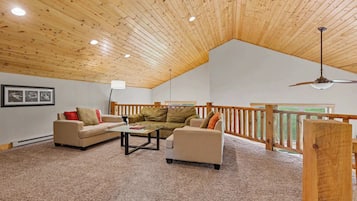A spacious living room with a high wooden ceiling, a glass coffee table, two sofas, and a wooden railing overlooking a view.