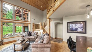 Ein geräumiges Wohnzimmer mit hoher Decke, großen Fenstern, einer Holztreppe und Blick ins Freie.