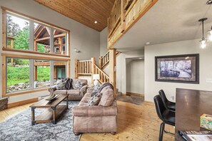A spacious living room with a high ceiling, large windows, a wooden staircase, and a view of the outdoors.