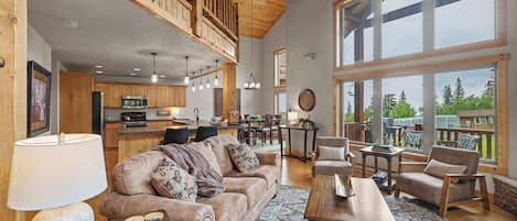A spacious living area with a high ceiling, wooden beams, a large window, and a kitchen in the background.