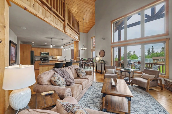 A spacious living area with a high ceiling, wooden beams, a large window, and a kitchen in the background.