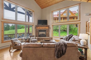 A spacious living room with a stone fireplace, large windows, and a view of a wooden cabin.