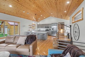 A spacious living area with a wooden ceiling, a kitchen with modern appliances, and a large window overlooking a wooden structure outside. - Kussy Slopeside Chalet at Terry Peak (Lead)