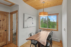 Et spiseområde med et træbord og stole, en hængende lampe, et vindue med udsigt til et skovområde og en dør med træpaneler.