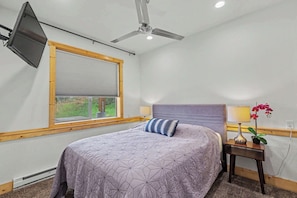 Ein Schlafzimmer mit einem Bett, einem Deckenventilator, einem Fenster mit Jalousien, einem Nachttisch mit Lampe und Pflanze sowie einem an der Wand montierten Fernseher.
