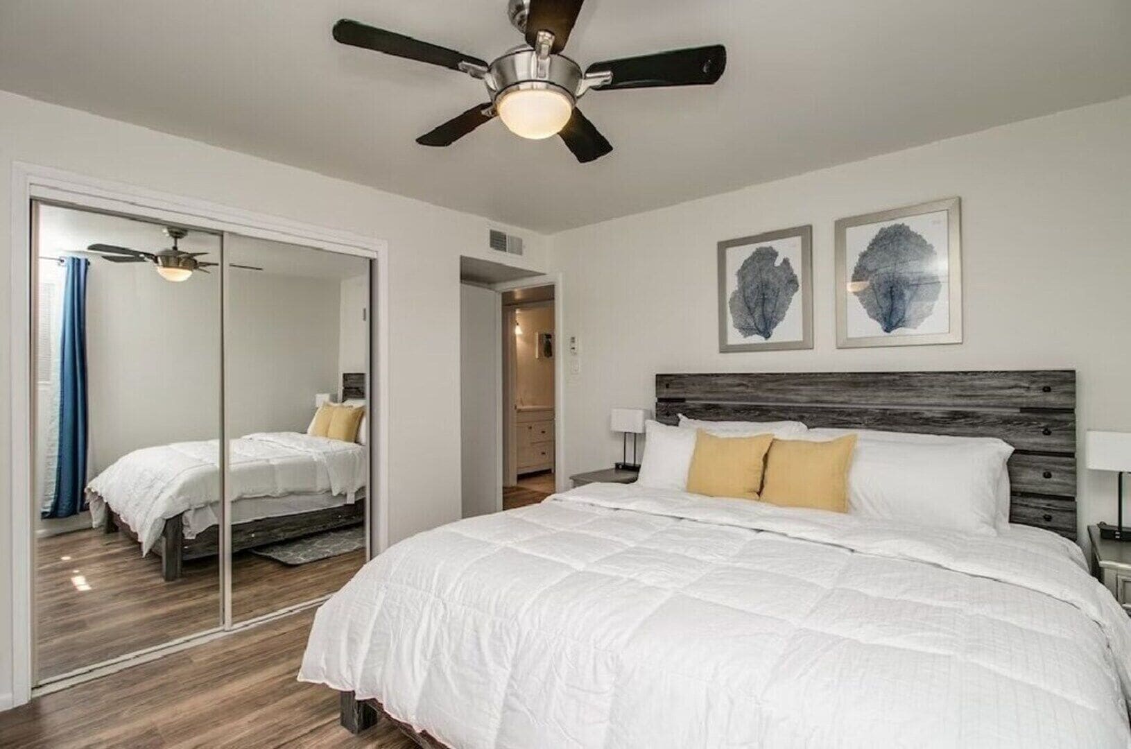 Quarto com uma cama grande, ventilador de teto, espelho e duas obras de arte penduradas na parede.