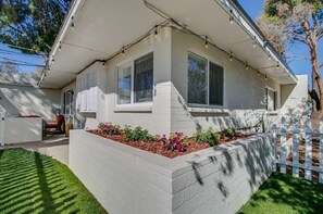 Ein eingeschossiges Haus mit einer Veranda, einem kleinen Garten mit Blumen und einem weißen Zaun.