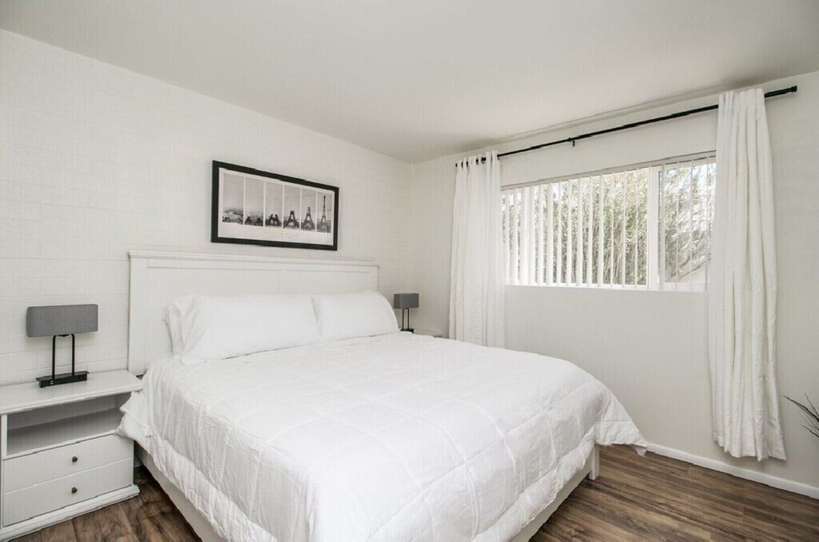 A white bedroom with a bed, a nightstand, a lamp, a window with curtains, and a framed picture on the wall.