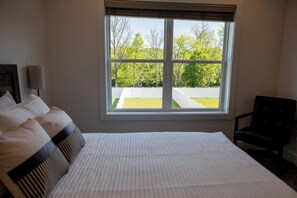 A bedroom with a large bed, a chair, and a window with a view of a white fence and trees. - Modern Pigeon River Retreat | Pet Friendly! (Sevierville)