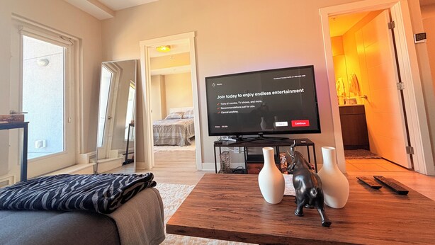 A hotel room with a bed, a TV, a wooden table with vases, and a bathroom visible through an open door. - Condo Downtown (Beltline) w/ Underground Parking (Calgary)