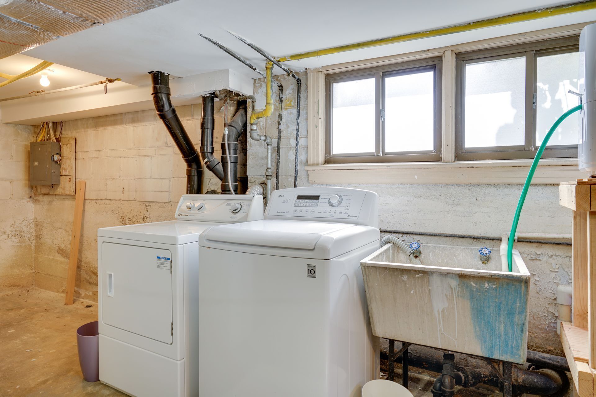 A laundry room with two white washing machines, a large window, and exposed plumbing.