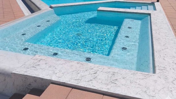 Uma banheira de hidromassagem com formato quadrado e borda de mármore, cercada por um deck revestido com azulejos.
