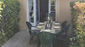 Pátio externo com mesa posta para quatro pessoas, rodeado por cadeiras, sob um guarda-sol branco.