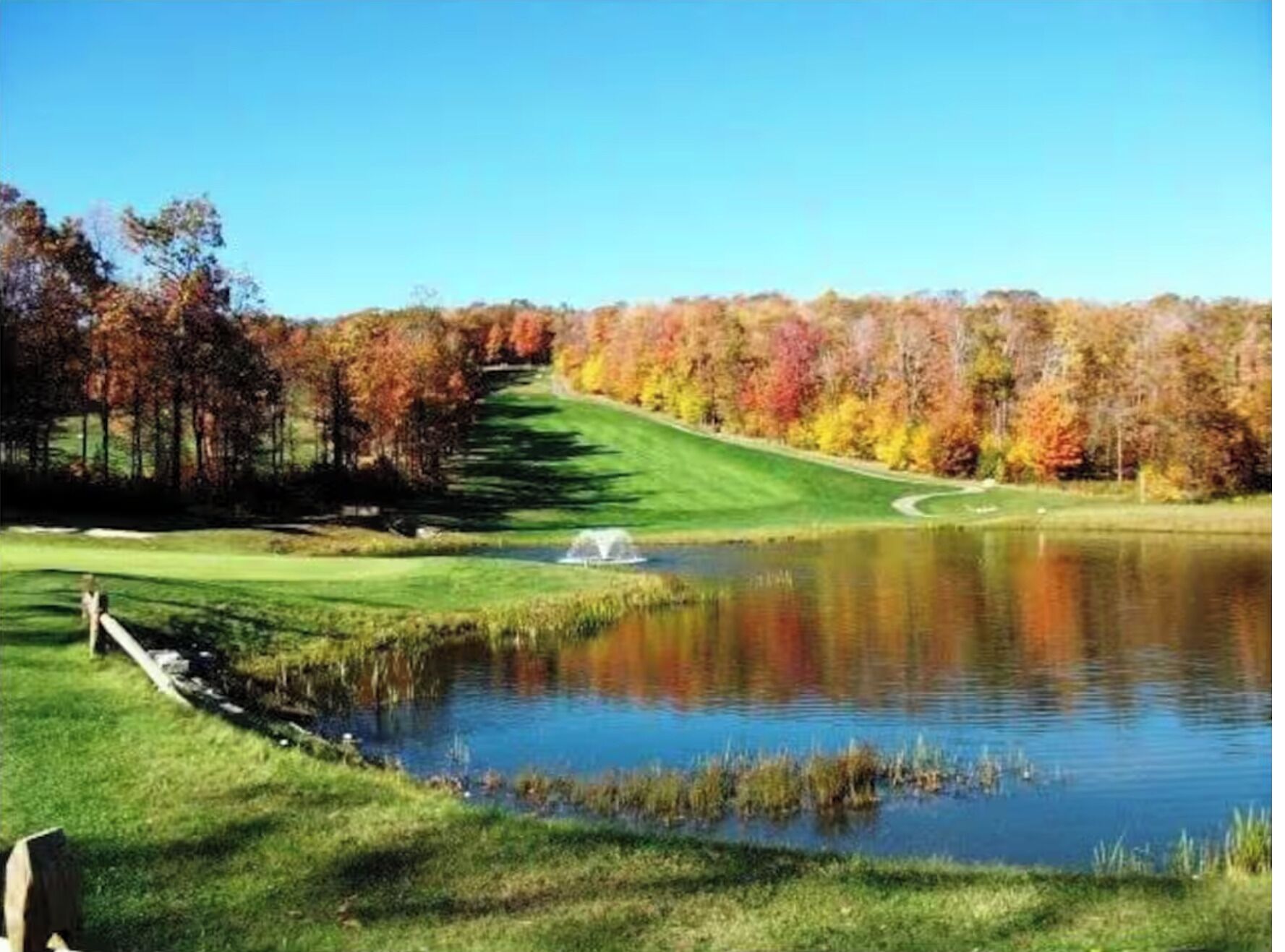 A golf course with a pond, trees in autumn colors, and a clear sky.