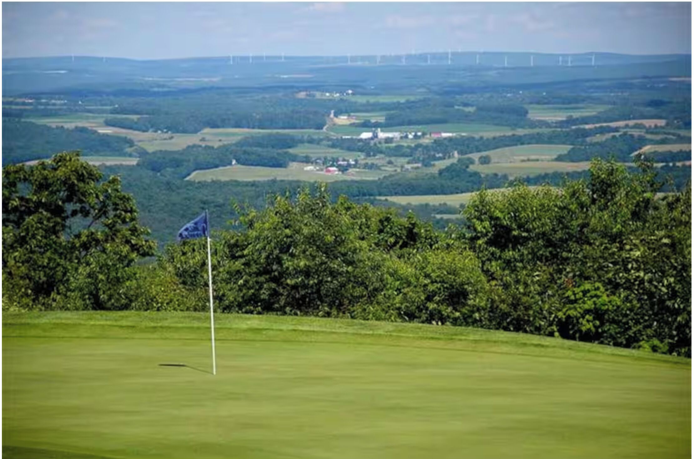 A golf course with a flagstick, surrounded by trees and rolling hills.