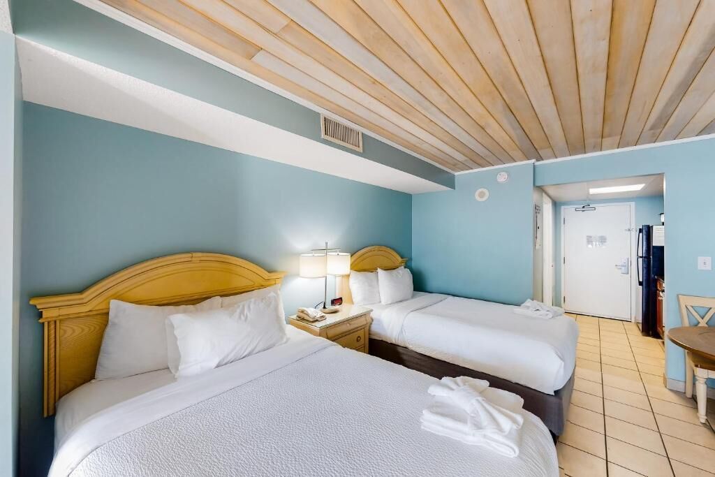 A hotel room with two beds, a wooden ceiling, a small table, and a door leading to a bathroom.