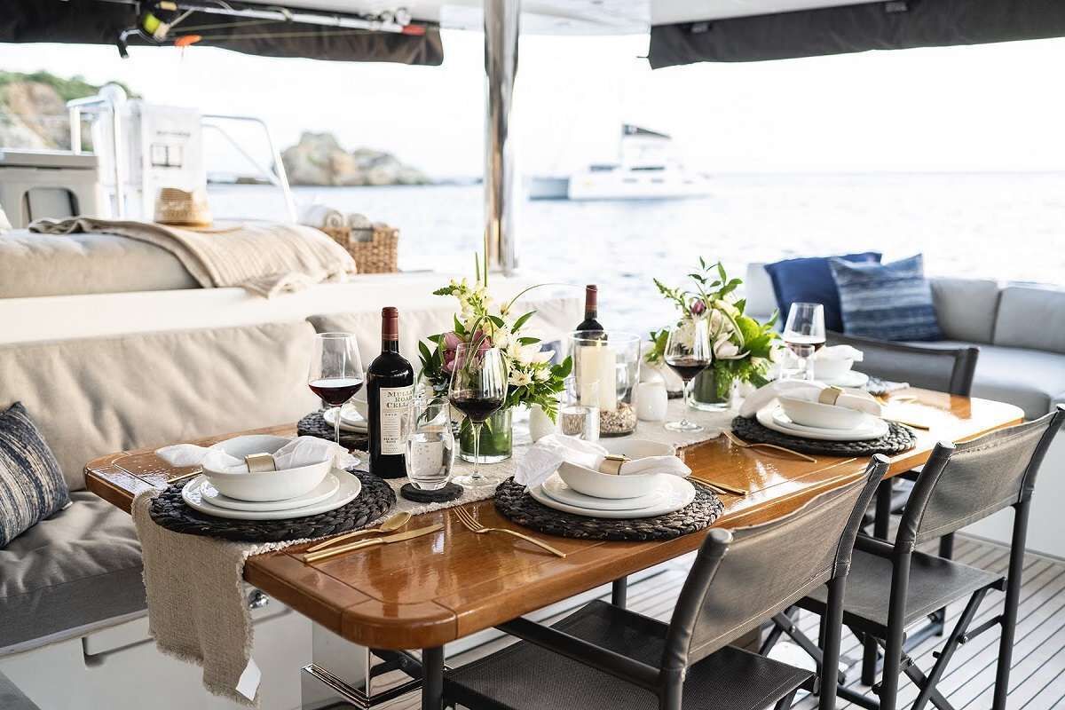 A dining table set with plates, glasses, and a bottle of wine, with a view of the sea and boats in the background.