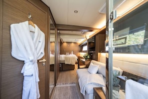 A modern interior with a wooden wardrobe, a white bathrobe, a bed, a table with a lamp, and a window with a view. - Floating Villa: All-Inclusive Yacht Vacation with Captain & Chef - 8 Guests (Tortola)