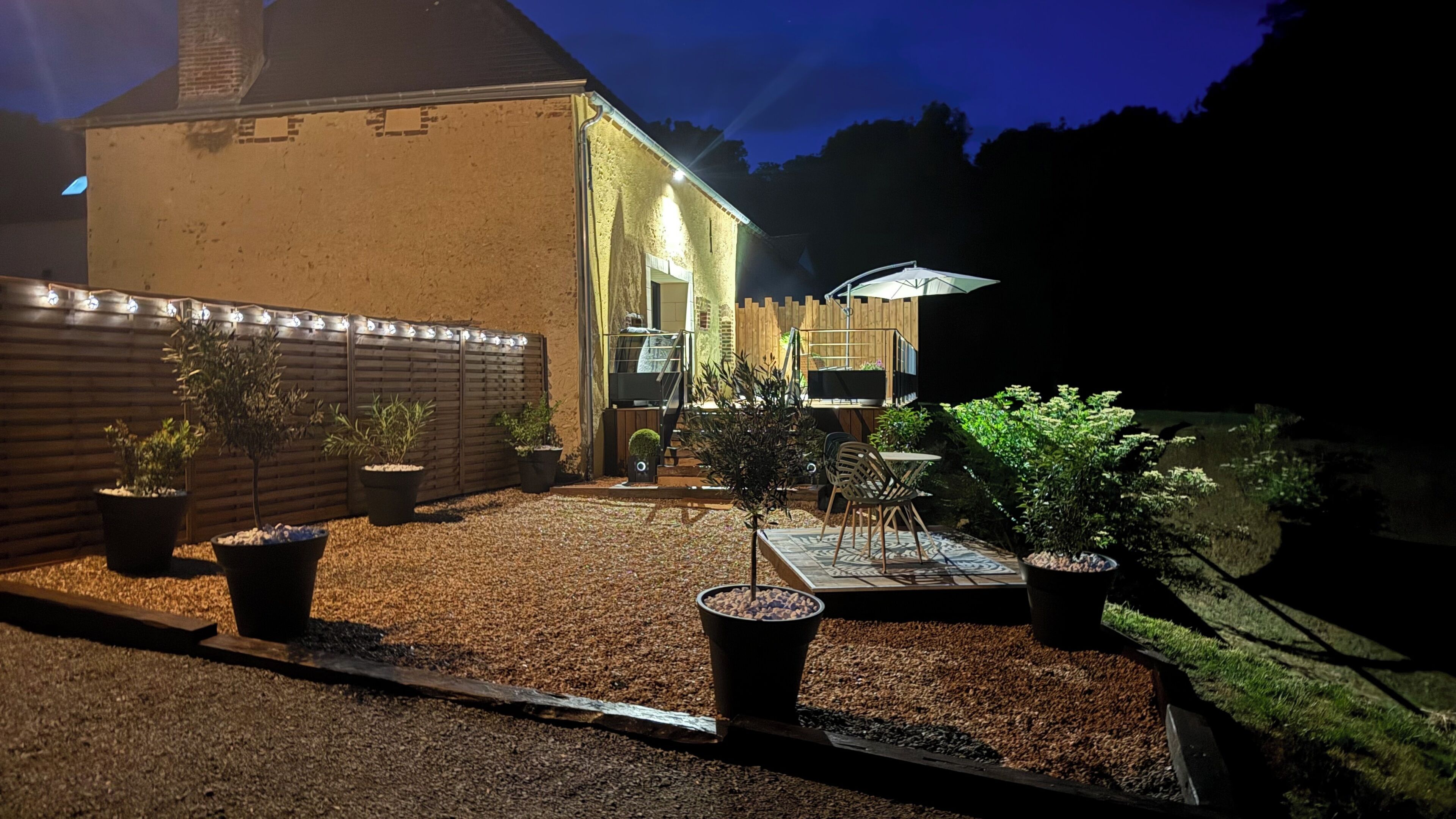 Un espace extérieur bien éclairé, aménagé en patio, avec un chemin de gravier, des plantes en pot, une clôture en bois et un bâtiment dont on aperçoit la porte et les fenêtres.