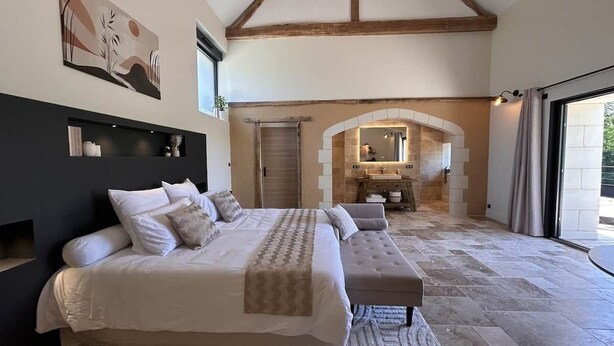 Un dormitorio con una cama grande, un banco, un baño con espejo y lavamanos, y un arco que da a un espacio exterior.