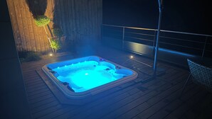Un jacuzzi en una terraza de madera con barandilla metálica y una pared al fondo.