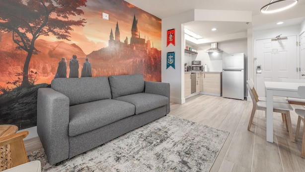 A modern living area with a grey sofa, a dining table with chairs, a kitchen with a fridge, and a wall mural. - Harry Potter Suite near Epic & Universal (Orlando)