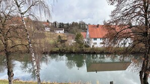 Ein ruhiger See mit Spiegelungen von Gebäuden und Bäumen, umgeben von grünen Wiesen und einer Straße.