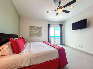 Habitació d'hotel amb un llit, un ventilador de sostre, una televisió, un quadre i vista a l'oceà per una porta corredissa.