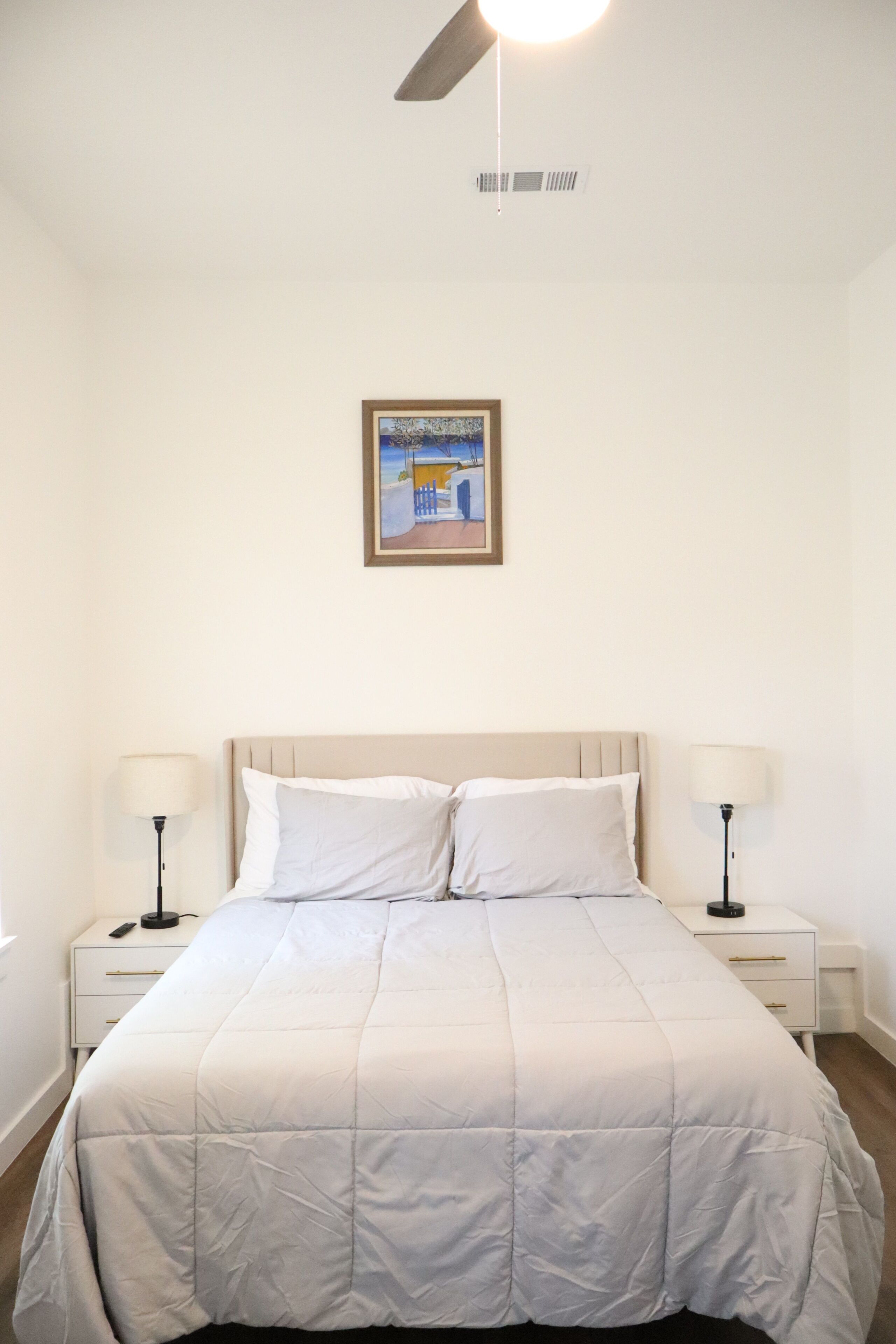 A bedroom with a bed, two bedside tables with lamps, a ceiling fan, and a framed picture on the wall.