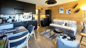Ein kompakter Wohnbereich mit Küchenzeile, einer Couch mit einem Ski-Kissen, einem Essbereich mit Tisch und Stühlen sowie einer dekorativen Lampe.