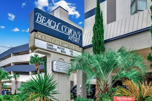 Ein mehrstöckiges Gebäude mit einem auffälligen Schild für das Beach Colony Resort, inklusive Annehmlichkeiten wie einem Café, einer Lounge und einem Verkaufsbüro für Apartments mit Meerblick.