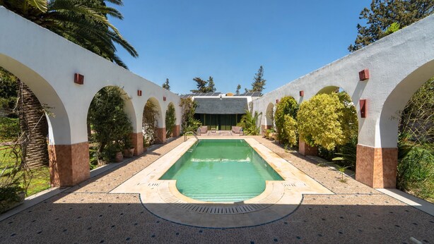 Une piscine dotée d’une terrasse carrelée, entourée de murs blancs percés d’arcades et agrémentée de verdure.