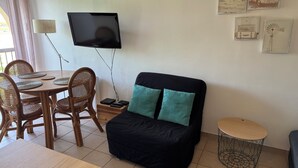 Een kamer met een aan de muur bevestigde televisie, een zwarte bank met blauwgroene kussens, een eettafel met geweven onderzetters, rieten stoelen en een kleine ronde tafel met een draadframe.